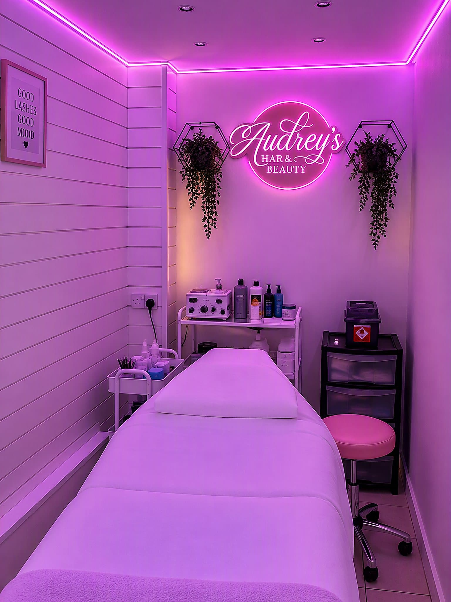 Inside Audrey's pink-lit treatment room — the white treatment bed beneath the glowing circular 'Audrey's Hair & Beauty' neon sign